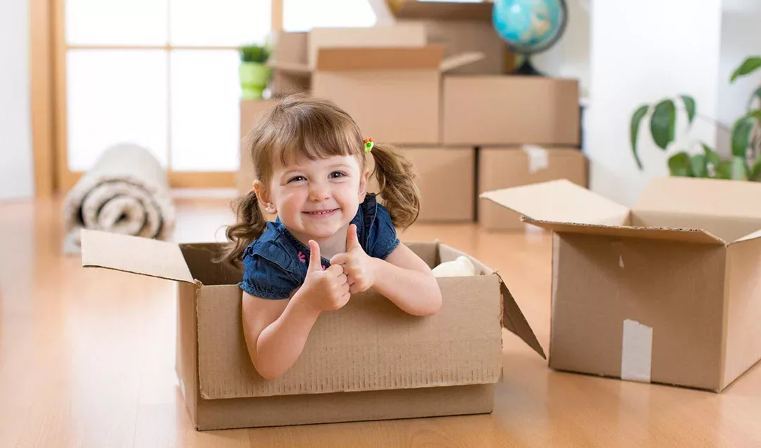 A toddler sitting in a moving box with a thumbs up.