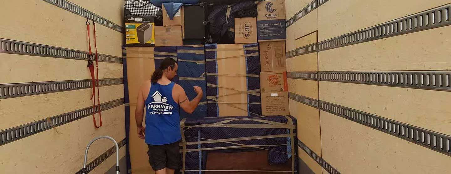 A person stands inside a moving truck filled with neatly arranged boxes and furniture, some covered with blankets for protection. The walls of the truck have metal rails and straps to secure the items—excellent moving tips on display.