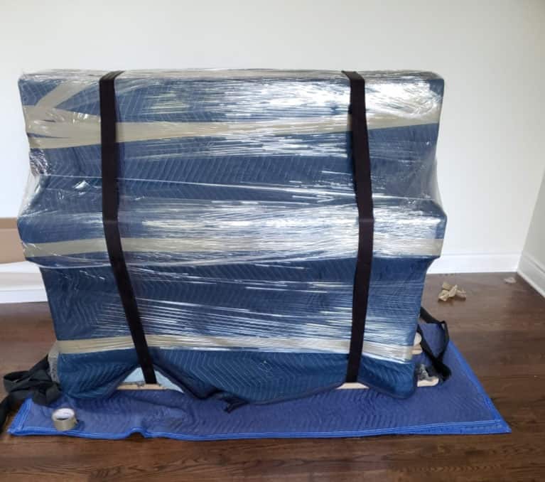 Piano wrapped in protective padding and plastic wrap, secured with straps by Ottawa piano movers, sitting on a blue blanket on a wooden floor.