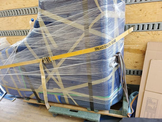 A piano wrapped in protective padding and plastic wrap, secured with straps on a wooden pallet inside a moving truck, prepared by Ottawa piano movers.