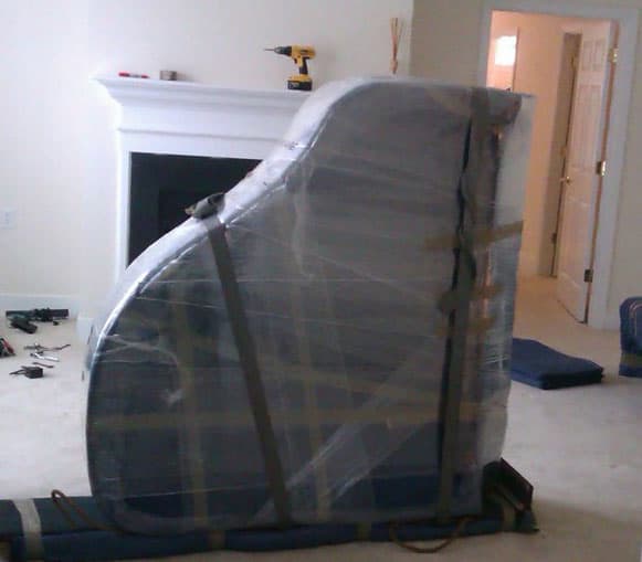 Wrapped grand piano secured with straps in a room with a fireplace and a drill on the mantel, expertly handled by Ottawa piano movers.