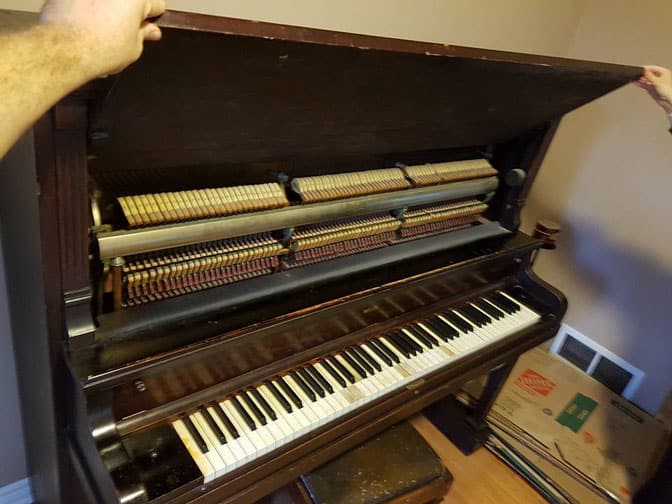 Two people holding the lid open to show an upright piano's internal mechanical components and keyboard in a room with packed boxes, likely prepped by Ottawa Piano Movers.