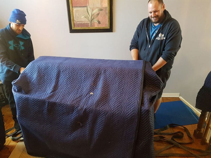 Two Ottawa piano movers covering and lifting a large piano draped with a padded blue moving blanket.