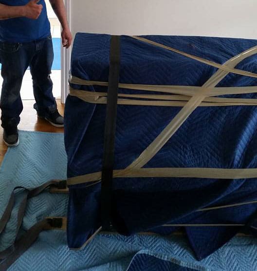 Mover standing beside a piano wrapped in a blue moving blanket secured with beige tape and straps.