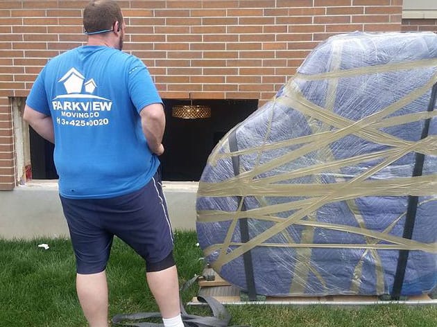 Mover standing next to a large, padded piano wrapped in tape outside a building during a move.