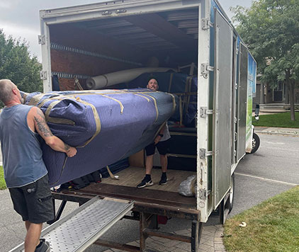 Two men load a large, wrapped item into a moving truck parked on a residential street.